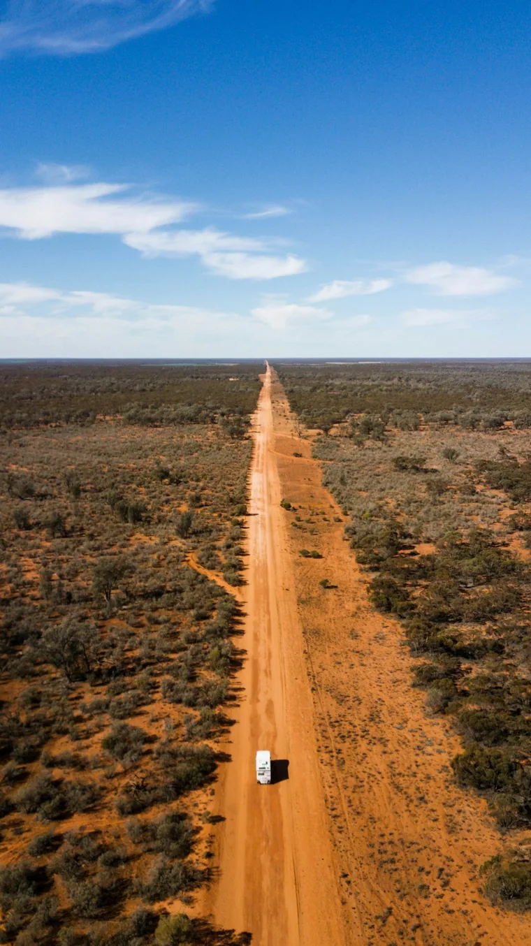 A motorhome driving along a long dirt road through dry scrub.